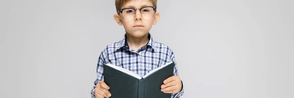 Un niño encantador con una camisa con incrustaciones y pantalones vaqueros ligeros se levanta sobre un fondo gris. El chico tiene un libro en sus manos. Niño con gafas —  Fotos de Stock