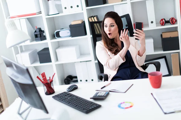 Egy fiatal lány ül a számítógépen az Office asztali, tartja a telefont, és kinyitotta a száját, az öröm. — Stock Fotó