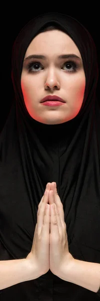 Retrato de una hermosa joven musulmana seria usando hiyab negro con las manos cerca de su cara como concepto de oración sobre fondo negro — Foto de Stock