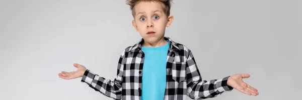 A handsome boy in a plaid shirt, blue shirt and jeans stands on a gray background. The boy spread his hands in both directions — Stock Photo, Image