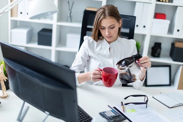 Een jong meisje zitten achter een computer bureau in het kantoor en giet zijn koffie. — Stockfoto