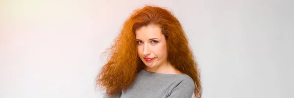 Charmante jonge meisje met rood haar. Een jong meisje in een grijze trui. Jong meisje op een grijze achtergrond. Jong meisje haar armen gevouwen — Stockfoto