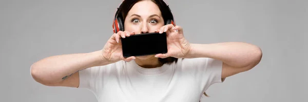 Retrato de emocional bonito modelo feliz plus size em fones de ouvido segurando telefone celular atrás de sua boca ouvindo música — Fotografia de Stock
