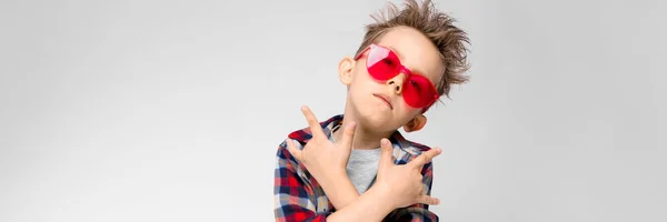 Um menino bonito em uma camisa xadrez, camisa cinza e jeans fica em um fundo cinza. Um rapaz de óculos de sol vermelhos. O menino mostra um bode roqueiro . — Fotografia de Stock