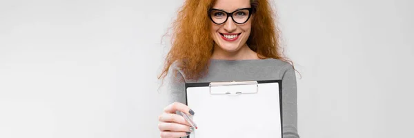 Retrato de hermosa pelirroja feliz sonriente joven mujer de negocios en ropa gris en gafas sujetando portapapeles sobre fondo gris —  Fotos de Stock