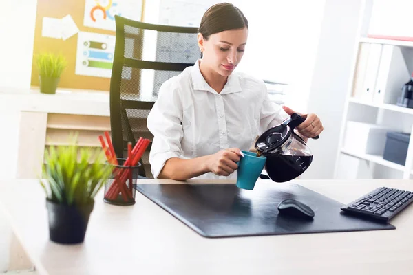 Una joven sentada en una mesa en la oficina y vierte café en una taza . — Foto de Stock