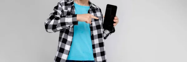 Um menino bonito em uma camisa xadrez, camisa azul e jeans fica em um fundo cinza. O rapaz está a segurar um telefone. — Fotografia de Stock