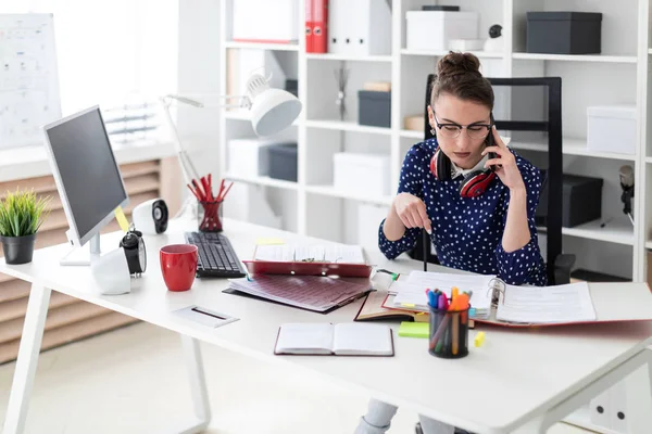 Una joven con gafas se sienta en la oficina en un escritorio de la computadora y está hablando por teléfono. Ante ella en la mesa yacen carpetas con documentos . — Foto de Stock
