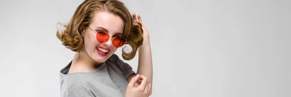 Encantadora joven con una camiseta gris sobre un fondo gris. Chica feliz en gafas rojas. La chica está medio despierta. —  Fotos de Stock