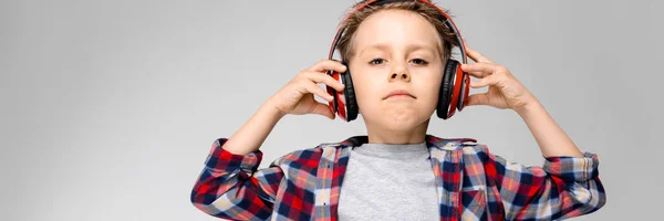 Un chico guapo con una camisa a cuadros, camisa gris y jeans está parado sobre un fondo gris. Un chico con auriculares rojos. El chico sostiene sus auriculares. . — Foto de Stock