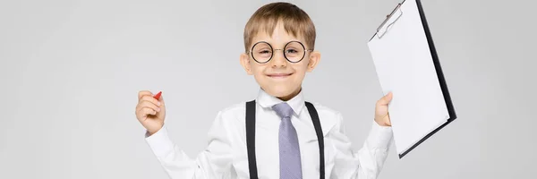 Um menino encantador com uma camisa branca, suspensórios, uma gravata e jeans leves fica em um fundo cinza. O menino segura uma caneta e lençóis para anotações — Fotografia de Stock