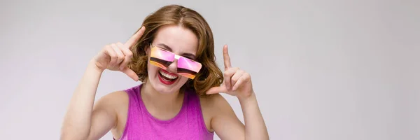 Encantadora joven en vestido rosa sobre fondo gris. Chica alegre en gafas cuadradas. La chica señala los dedos en la parte superior —  Fotos de Stock