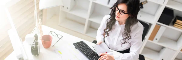 Una giovane ragazza sta lavorando al computer in ufficio e tiene in mano una penna e un quaderno . — Foto Stock