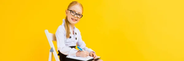 Menina com cabelo vermelho em um fundo amarelo. Uma menina encantadora em óculos transparentes senta-se em uma cadeira branca e faz anotações em um caderno . — Fotografia de Stock