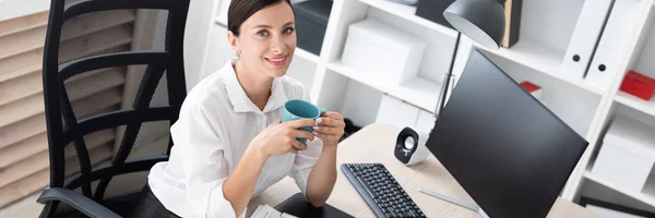 A young girl sitting in the office at the computer and holding a Cup. — Stock Photo, Image