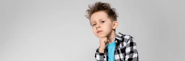 Um menino bonito em uma camisa xadrez, camisa azul e jeans fica em um fundo cinza. O menino sustenta o queixo com a mão — Fotografia de Stock