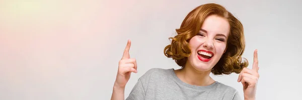 Charming young girl in a gray T-shirt on a gray background. The girl points fingers upward — Stock Photo, Image