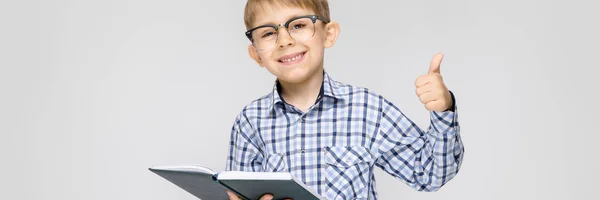 Um menino encantador com uma camisa embutida e jeans leves fica em um fundo cinza. O rapaz tem um livro nas mãos. Menino com óculos — Fotografia de Stock