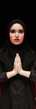 Güzel ciddi genç Müslüman kadın kavramı siyah arka plan üzerine dua olarak yüzünü yakınındaki elleriyle siyah hijab giyen portresi