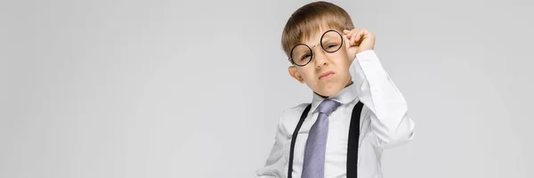 Un niño encantador con una camisa blanca, tirantes, corbata y pantalones vaqueros ligeros se levanta sobre un fondo gris. el niño levantó sus gafas —  Fotos de Stock