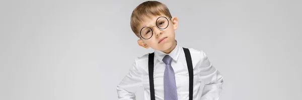 Un garçon charmant dans une chemise blanche, des bretelles, une cravate et un jean clair se dresse sur un fond gris. le garçon à lunettes pencha la tête sur le côté — Photo