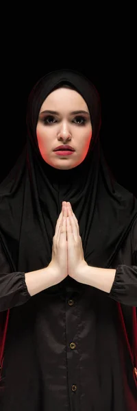 Porträt einer schönen, ernsthaften jungen Muslimin, die einen schwarzen Hijab trägt, die Hände vor dem Gesicht als Gebetskonzept auf schwarzem Hintergrund — Stockfoto