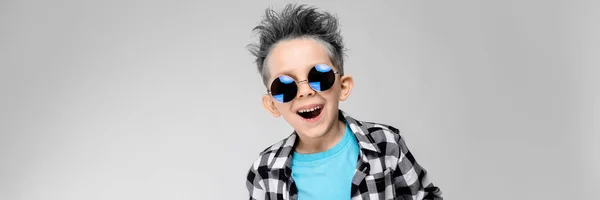 Een knappe jongen in een plaid shirt, blauw shirt en spijkerbroek staat op een grijze achtergrond. De jongen is een ronde bril. De jongen glimlacht. De jongen heeft een gewone haarkleur — Stockfoto