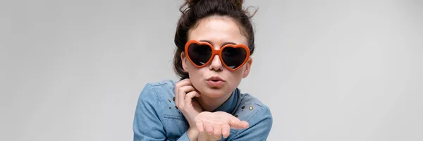 Junges brünettes Mädchen mit Brille in Form eines Herzens. Die Haare sind zu einem Dutt zusammengebunden. das Mädchen schickt einen Luftkuss. — Stockfoto