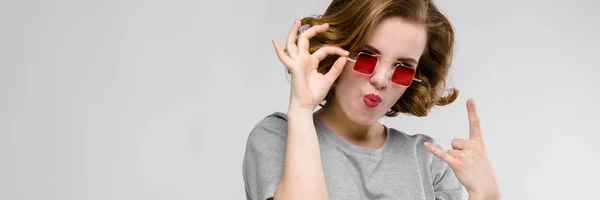 Encantadora joven con una camiseta gris sobre un fondo gris. Chica con gafas rojas de ojos cuadrados. La chica muestra una cabra rockera —  Fotos de Stock