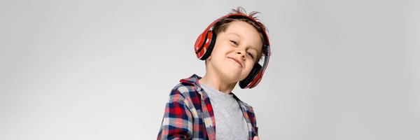 Um menino bonito em uma camisa xadrez, camisa cinza e jeans fica em um fundo cinza. Um menino de fones de ouvido vermelhos ouve rap . — Fotografia de Stock