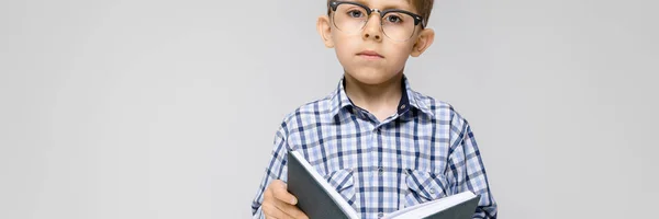 En charmig pojke med en inläggningar skjorta och ljus jeans står på en grå bakgrund. Pojken håller en bok i händerna. Pojke med glasögon — Stockfoto