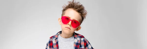 Un chico guapo con una camisa a cuadros, camisa gris y jeans está parado sobre un fondo gris. Niño en gafas de sol rojas — Foto de Stock