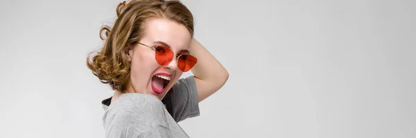 Menina encantadora em uma camiseta cinza em um fundo cinza. Menina feliz em óculos vermelhos. A rapariga está de pé meio acordada — Fotografia de Stock