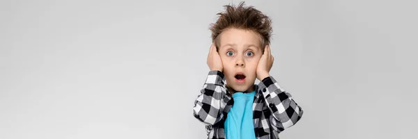 Um menino bonito em uma camisa xadrez, camisa azul e jeans fica em um fundo cinza. O menino abriu a boca e tapou os ouvidos com as mãos — Fotografia de Stock