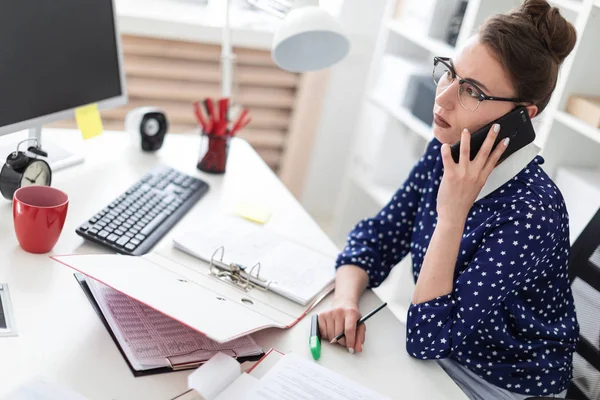 Een jong meisje in glazen zit op kantoor op een computerbureau en is praten over de telefoon. Voor haar op tafel liggen mappen met documenten. — Stockfoto