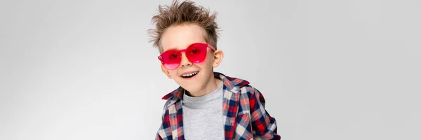 Ένα όμορφο αγόρι σε ένα καρό πουκάμισο, γκρι πουκάμισο και τζιν στέκεται σε γκρι φόντο. Ένα αγόρι σε κόκκινα γυαλιά ηλίου. Το αγόρι εξάπλωση τα χέρια του προς τις δύο κατευθύνσεις — Φωτογραφία Αρχείου