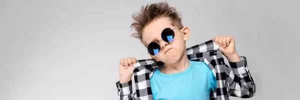 A handsome boy in a plaid shirt, blue shirt and jeans stands on a gray background. The boy is wearing round glasses. Redhead boy holding his fingers collar shirt — Stock Photo, Image