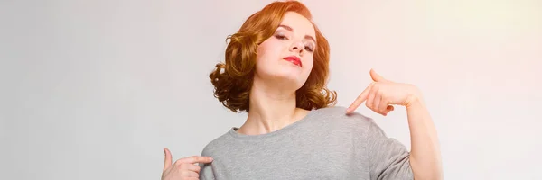 Affascinante ragazza in una t-shirt grigia su uno sfondo grigio. La ragazza punta il dito contro se stessa — Foto Stock