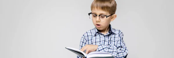 Un garçon charmant avec une chemise incrustée et un jean clair se dresse sur un fond gris. Le garçon tient un livre entre ses mains. Garçon avec des lunettes — Photo