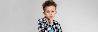 Yakışıklı bir çocuk bir ekose gömlek, mavi tişört ve kot gri bir arka plan üzerinde duruyor. Çocuk çenesini eliyle destekler.