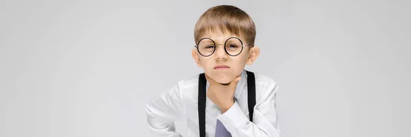 Un niño encantador con una camisa blanca, tirantes, corbata y pantalones vaqueros ligeros se levanta sobre un fondo gris. un chico con gafas está sosteniendo una mano cerca de su cara — Foto de Stock