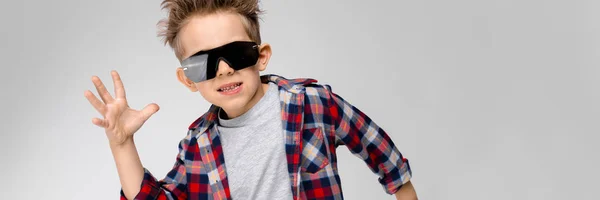 Yakışıklı bir çocuk bir ekose gömlek, gri gömlek ve pantolon gri bir arka plan üzerinde duruyor. Siyah güneş gözlüklü çocuk. Çocuk ellerini yanlara yaymak. — Stok fotoğraf