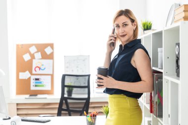 Ofiste telefonda konuşuyor ve bir bardak kahve tutan genç bir kız.