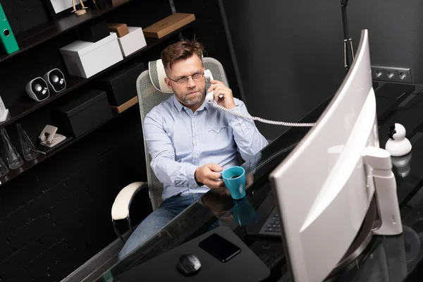 Man leaned back in his chair, drinking coffee and talking on landline — Stock Photo, Image