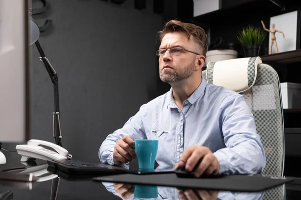 Деловой человек в очках, работающий в офисе за компьютерным столом и пьющий кофе из маленькой чашки — стоковое фото