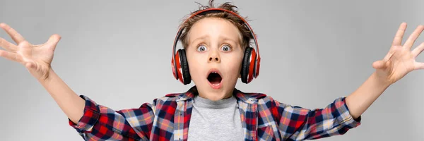 Een knappe jongen in een plaid shirt, grijs shirt en spijkerbroek staat op een grijze achtergrond. Een jongen in rode hoofdtelefoon. De jongen trekt zijn handen naar voren. — Stockfoto
