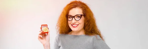 Портрет красивой рыжеволосой счастливой улыбающейся молодой женщины в серой одежде в очках, показывающей биткойн как концепцию криптовалюты на сером фоне — стоковое фото