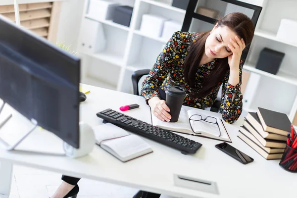 Una joven sentada en la oficina en la mesa de la computadora y sosteniendo un vaso de café . — Foto de Stock