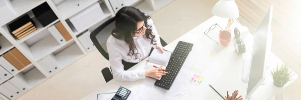 Genç bir kız ofiste bilgisayar çalışma ve bir kalem ve defter elinde tutan.