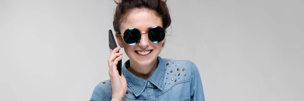 Menina morena nova em óculos redondos. Os pêlos estão reunidos num pão. Menina com um telefone preto. A rapariga está a falar ao telefone. . — Fotografia de Stock